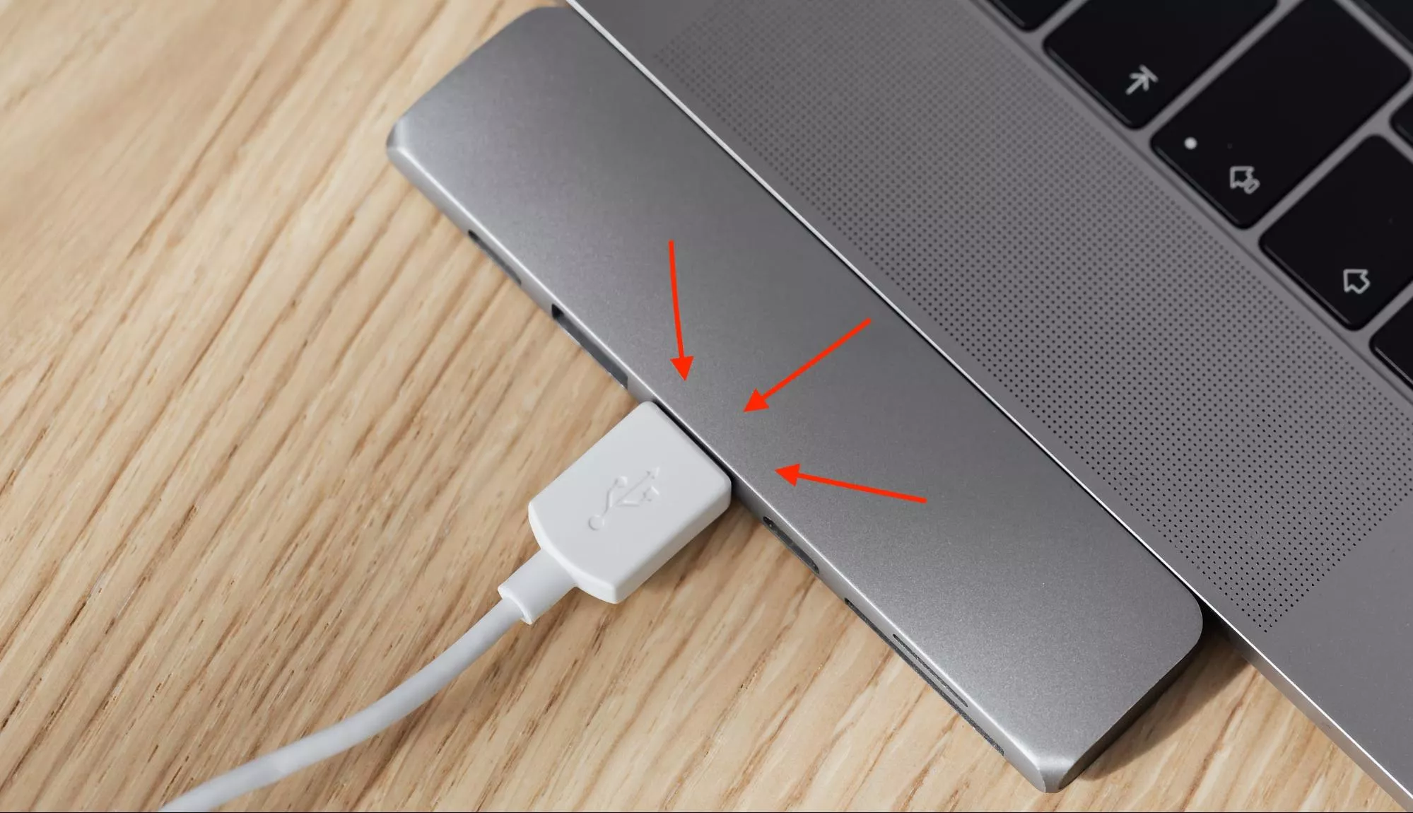 Foto de um cabo USB branco conectado a um dispositivo Mac sobre uma superfície de madeira. Três setas vermelhas apontam para o cabo USB indicando onde o leitor deve verificar.